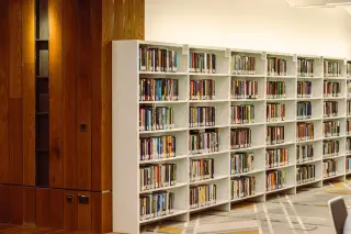 Ilustrasi: Deretan rak putih berisi buku-buku di dalam ruangan perpustakaan berdinding kayu.
