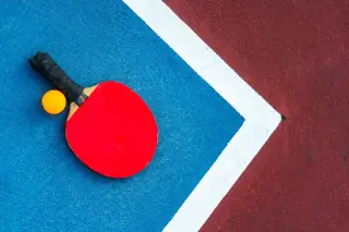 Ilustrasi: Bet tenis meja berwarna merah dan bola oranye terletak di atas lantai lapangan olahraga.