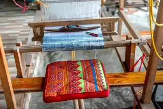 Ilustrasi: Alat tenun tradisional berbahan kayu dengan kain tenun dan bantal bermotif warna-warni.