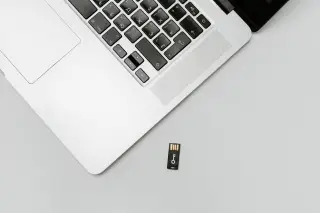 Ilustrasi: Sebagian laptop terbuka dan sebuah diska lepas USB kecil di atas permukaan abu-abu polos.