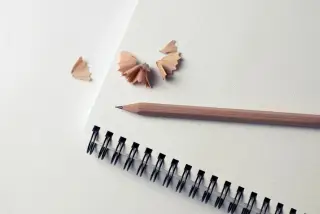 Ilustrasi: Sebuah pensil kayu dan serutan pensil di atas buku catatan bergaris titik.