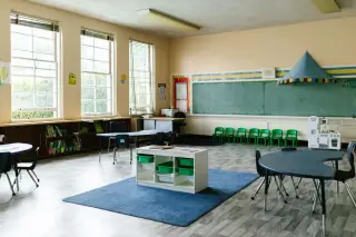 Ilustrasi: Ruang kelas prasekolah yang luas dan kosong dengan meja, kursi, serta papan tulis hijau.