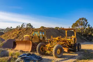 Ilustrasi: Dua alat berat buldoser dan grader kuning terparkir di area proyek tanah berbukit.