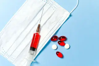Ilustrasi: Masker medis, jarum suntik berisi cairan merah, dan obat kapsul serta pil di atas latar biru.