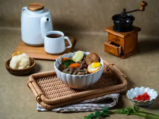 Ilustrasi: Semangkuk sup daging sapi dengan telur dan pelengkap disajikan di atas nampan rotan.