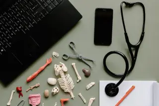 Ilustrasi: Stetoskop, laptop, ponsel, dan model miniatur anatomi tubuh di atas meja kerja.