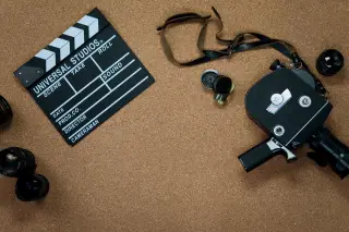 Ilustrasi: Papan klaper film, kamera video klasik, dan lensa kamera di atas latar belakang gabus cokelat.