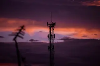 Ilustrasi: Siluet menara telekomunikasi berdiri tegak dengan latar langit senja berawan ungu dan jingga.