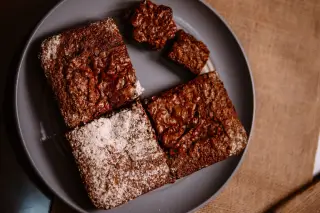 Ilustrasi: Potongan kue brownies cokelat ditaburi gula halus di atas piring bundar abu-abu.