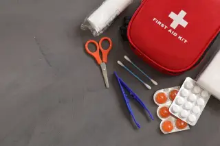 Ilustrasi: Kotak P3K merah beserta gunting, pinset, obat, dan perban di atas meja abu-abu.