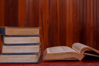 Ilustrasi: Tumpukan buku tebal dan sebuah buku terbuka di atas meja dengan latar dinding kayu.