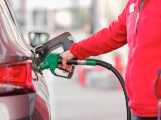 Ilustrasi: Seseorang berjaket merah mengisi bahan bakar ke tangki mobil menggunakan selang dispenser hijau.