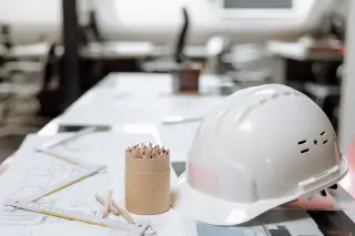 Ilustrasi: Helm proyek putih, pensil warna, dan cetak biru arsitektur di atas meja kerja kantor.