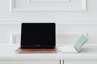 Ilustrasi: Sebuah laptop terbuka dan buku catatan kecil di atas meja putih dengan latar dinding minimalis.