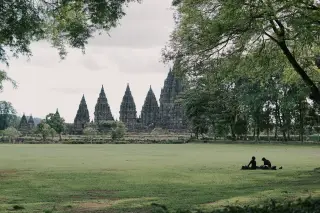 Ilustrasi: Pemandangan Candi Prambanan di kejauhan dengan dua orang duduk santai di lapangan rumput.