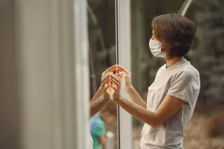 Ilustrasi: Seorang wanita bermasker berdiri menatap ke luar dari balik jendela kaca besar.