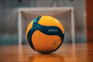 Ilustrasi: Sebuah bola voli berwarna kuning dan biru diletakkan di atas lantai lapangan olahraga dalam ruangan.
