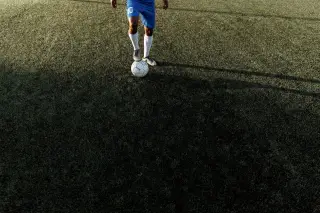 Ilustrasi: Kaki seorang pemain sepak bola menginjak bola di lapangan rumput dengan bayangan memanjang.