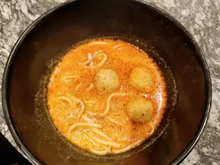 Ilustrasi: Semangkuk mi berkuah merah dengan tiga butir bakso di dalam mangkok hitam.