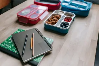 Ilustrasi: Buku catatan, pensil, dan wadah bekal makanan di atas meja kayu.