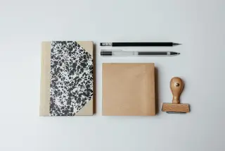 Ilustrasi: Buku catatan, pensil, pulpen, dan sebuah stempel kayu di atas permukaan putih.