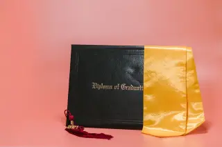 Ilustrasi: Map ijazah hitam dengan selempang kuning dan rumbai kelulusan di atas latar merah muda.