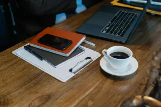 Ilustrasi: Laptop, cangkir kopi, ponsel, dan buku catatan di atas meja kayu.