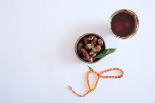 Ilustrasi: Secangkir teh, semangkuk kurma, dan seuntai tasbih dari sudut pandang atas di atas latar putih.