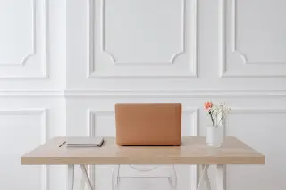 Ilustrasi: Sebuah laptop, buku catatan, dan vas bunga tertata rapi di atas meja kerja kayu dengan latar dinding putih.