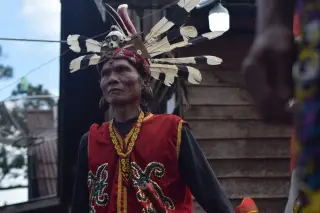 Ilustrasi: Pria mengenakan busana adat Dayak dan hiasan kepala berbulu di depan rumah kayu.