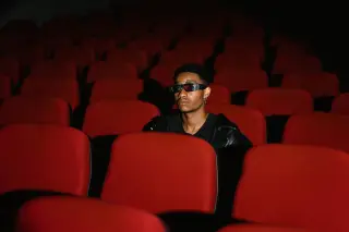Ilustrasi: Seorang pria berkacamata 3D duduk sendirian di antara deretan kursi bioskop merah.