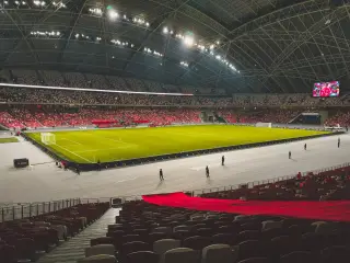 Ilustrasi: Pemandangan luas lapangan sepak bola dalam stadion tertutup dengan barisan tribun penonton.