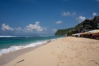 Ilustrasi: Suasana pantai berpasir putih dengan ombak laut, tebing, dan pengunjung di bawah terik matahari.