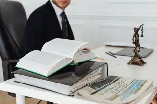 Ilustrasi: Buku hukum terbuka, berkas dokumen, dan patung keadilan di atas meja di depan pria berjas.