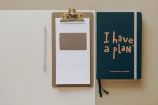Ilustrasi: Papan klip dengan kertas dan kartu cip di samping buku catatan bertuliskan 'I have a plan' serta sebuah pena.