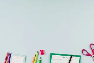 Ilustrasi: Alat tulis seperti pensil warna, gunting, dan buku catatan di atas latar biru polos.
