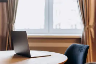 Ilustrasi: Sebuah laptop terbuka di atas meja bundar di dekat jendela dengan tirai.