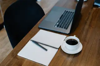 Ilustrasi: Buku catatan polos, pena, secangkir kopi, dan laptop di atas meja kayu.