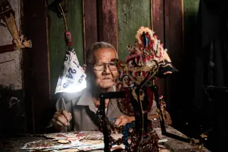 Ilustrasi: Seorang pria lanjut usia sedang membuat wayang kulit tradisional di bawah lampu meja.