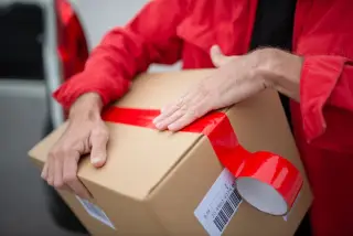 Ilustrasi: Seseorang berbaju merah merekatkan selotip merah pada kardus paket pengiriman.