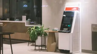 Ilustrasi: Sebuah mesin ATM berdiri di sudut ruangan dekat tanaman hias dan meja kecil.