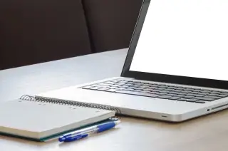 Ilustrasi: Laptop dengan layar putih kosong di samping buku catatan dan pena di atas meja.