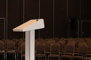 Ilustrasi: Sebuah podium putih dengan mikrofon berdiri di depan deretan kursi kosong di dalam ruangan.