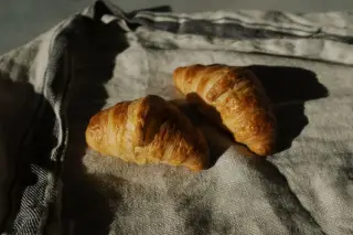 Ilustrasi: Dua buah roti croissant diletakkan di atas kain bermotif garis dengan pencahayaan alami.