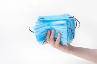 Ilustrasi: Satu tangan memegang tumpukan masker medis berwarna biru dengan latar belakang putih polos.