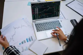 Ilustrasi: Seseorang menganalisis grafik saham di laptop dan lembaran data di atas meja kerja.