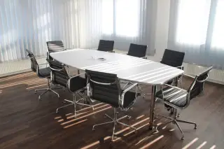 Ilustrasi: Meja rapat putih panjang dan kursi kantor hitam di dalam ruangan berlantai kayu.