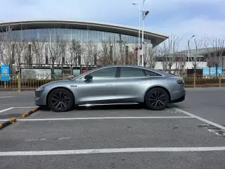 Ilustrasi: Mobil sedan modern berwarna abu-abu terparkir di area parkir luar ruangan depan gedung besar.