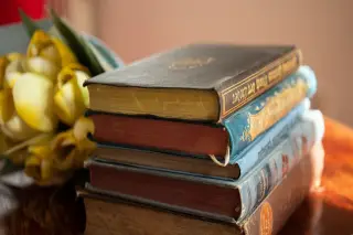 Ilustrasi: Tumpukan buku-buku antik bersampul tebal di samping bunga kuning di atas meja kayu.