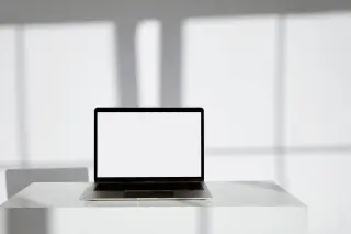 Ilustrasi: Sebuah laptop terbuka dengan layar putih di atas meja dengan latar belakang dinding putih berbayang.